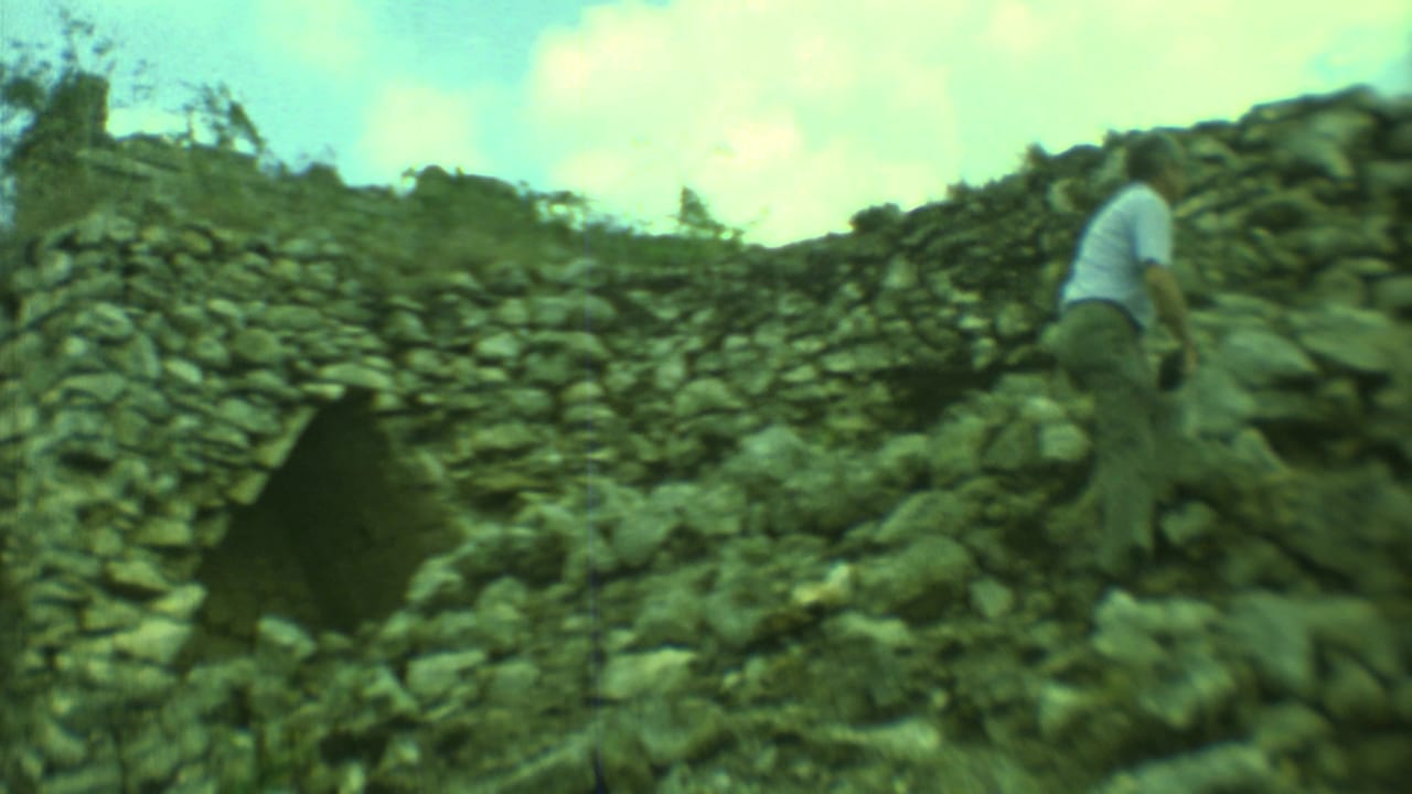 1973 Yucatan frame of an adult man climbing across rubble beside a weathered stone ruin wall under clouds.