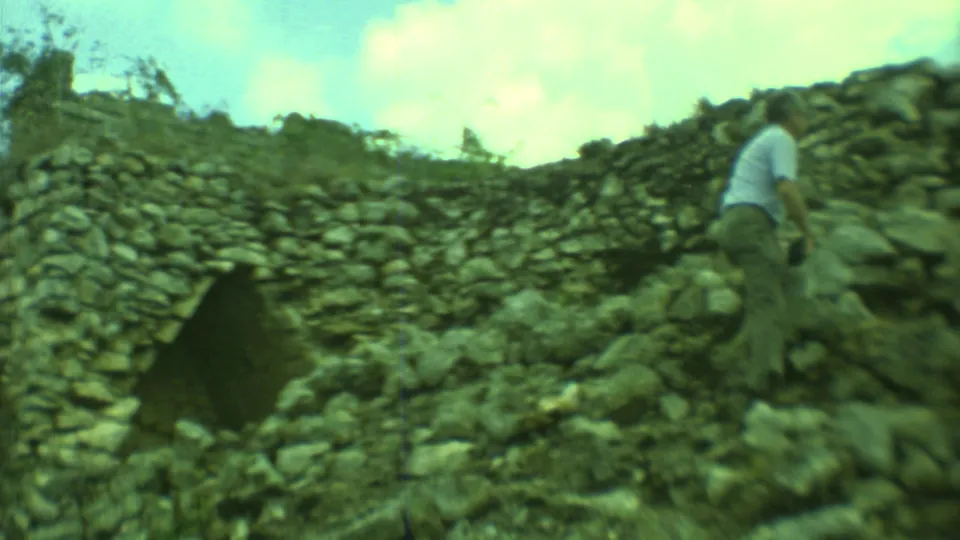 1973 Yucatan frame of an adult man climbing across rubble beside a weathered stone ruin wall under clouds.