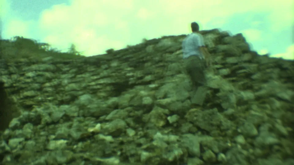 1973 Yucatan home-movie view of a man from behind climbing a rocky ruin slope with stonework and clouds beyond.
