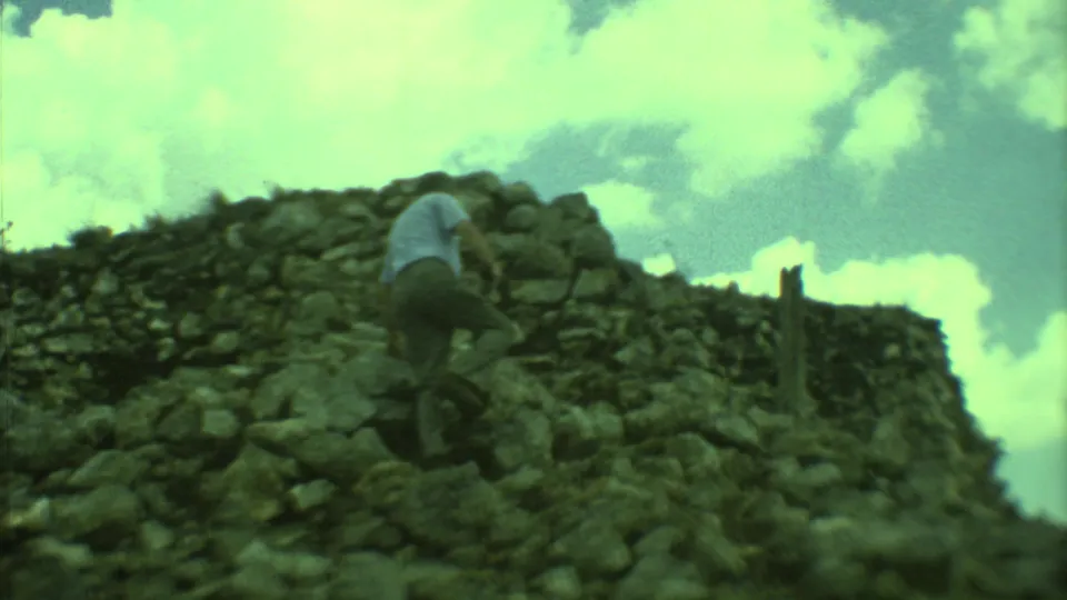 1973 Yucatan frame showing an adult man climbing near the upper stones of a weathered ruin against a bright cloudy sky.