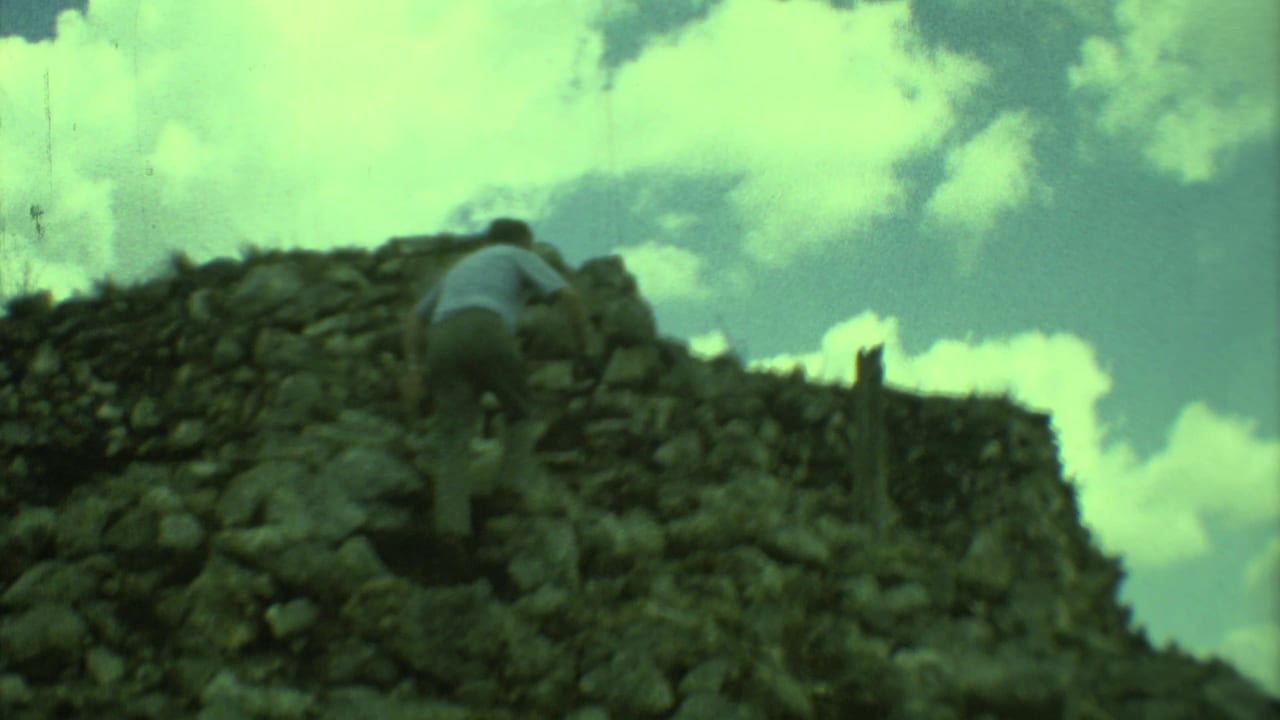 1973 8mm travel frame from Yucatan, Mexico, showing a man climbing a rough stone structure beneath bright clouds.