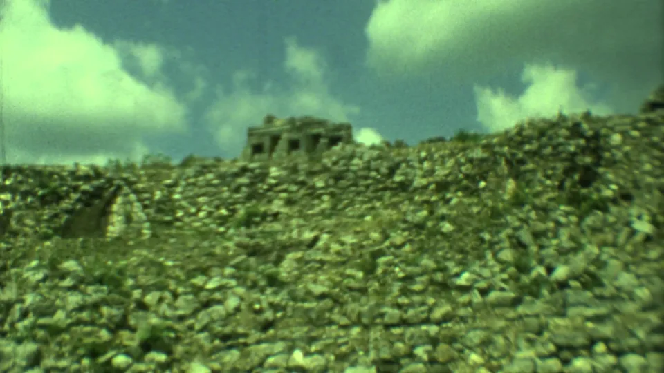 1973 Yucatan 8mm frame showing a rubble-strewn ruin mound and a distant rectangular stone structure beneath a bright sky.