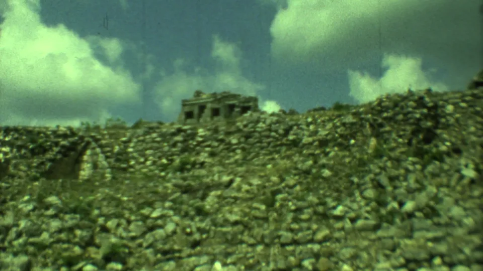 1973 Yucatan home-movie frame with a distant temple-like ruin beyond rubble-covered stonework and clouds.
