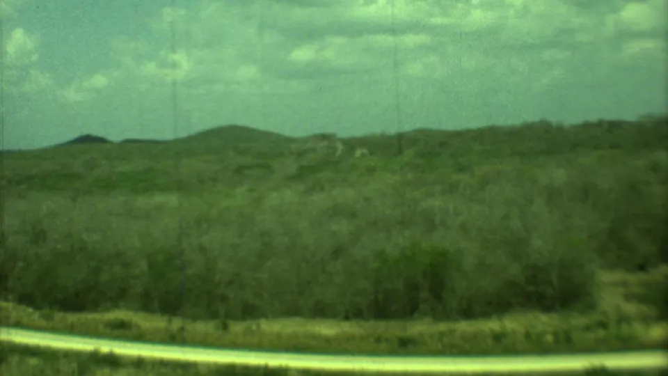 1973 Yucatan countryside view with rolling scrub-covered hills, a light road strip, and hazy 8mm film texture.