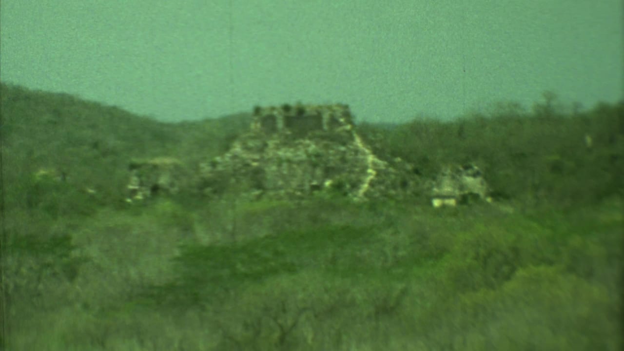 Weathered stone ruin partly hidden by green jungle and low hills.