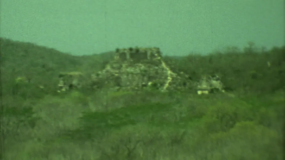1973 Yucatan home-movie frame showing a weathered stone ruin surrounded by dense vegetation and distant hills.