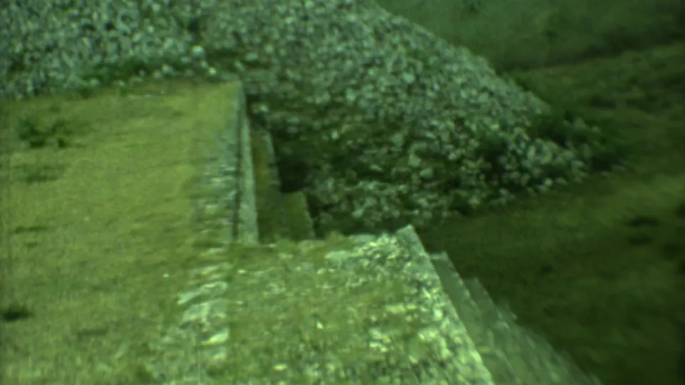 Green-tinted 8mm frame showing a Yucatan stone stairway with a nearby pile of rocks and open grassy ground.