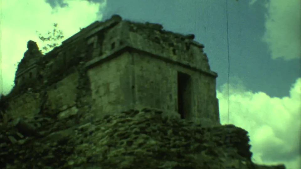 1973 Yucatan 8mm frame looking up at a weathered stone ruin with dark openings, rough masonry, sky, and clouds.