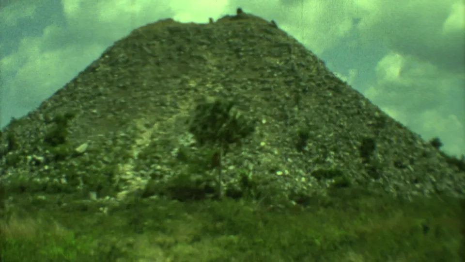 1973 Yucatan home-movie frame showing a large stone-covered mound or pyramid ruin with vegetation.