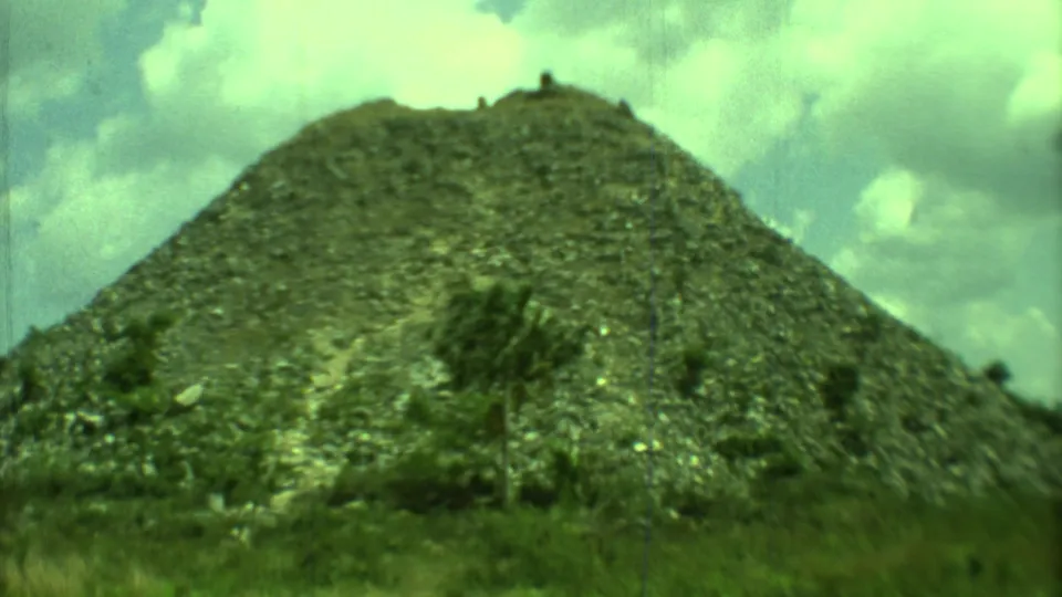 Vintage 8mm Yucatan frame of a sloped stone mound, low vegetation, and bright cloudy sky.