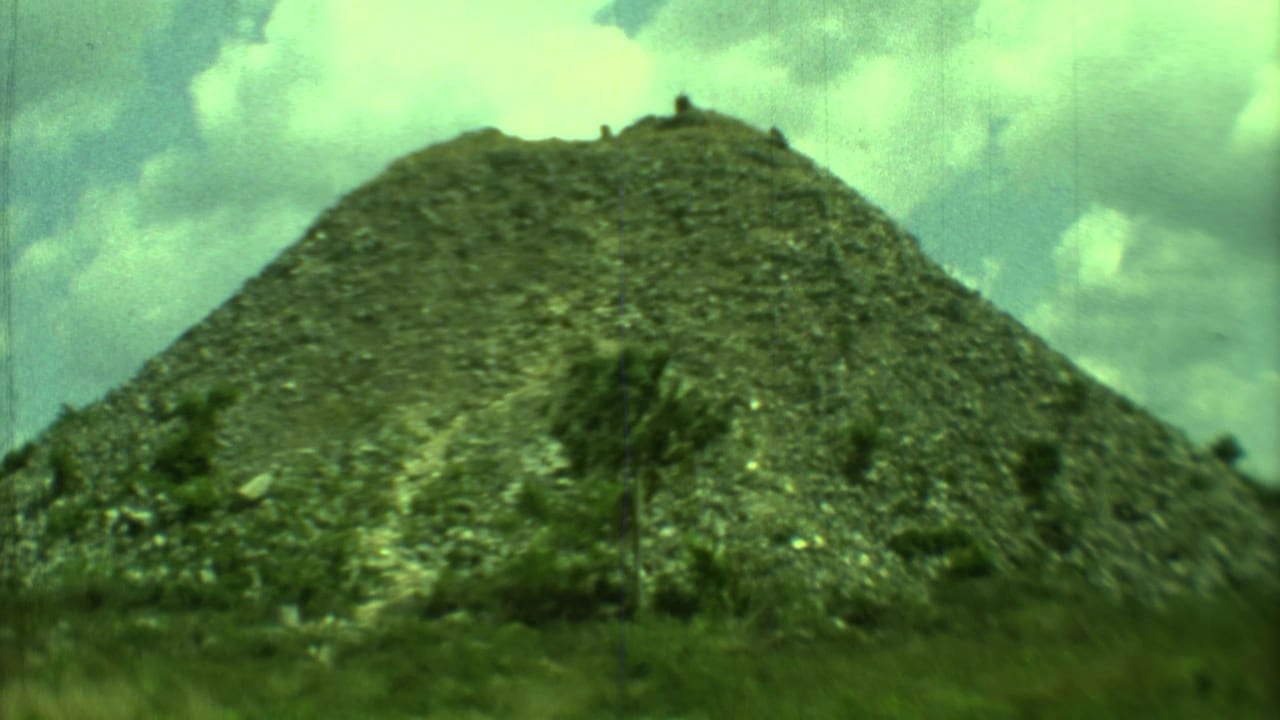 1973 Yucatan 8mm frame showing a steep vegetation-covered mound with scrubby foreground growth and clouds overhead.