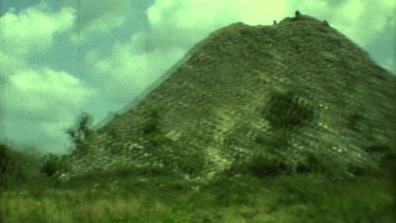 Vegetation-covered mound with rocky slopes and small trees in the foreground.