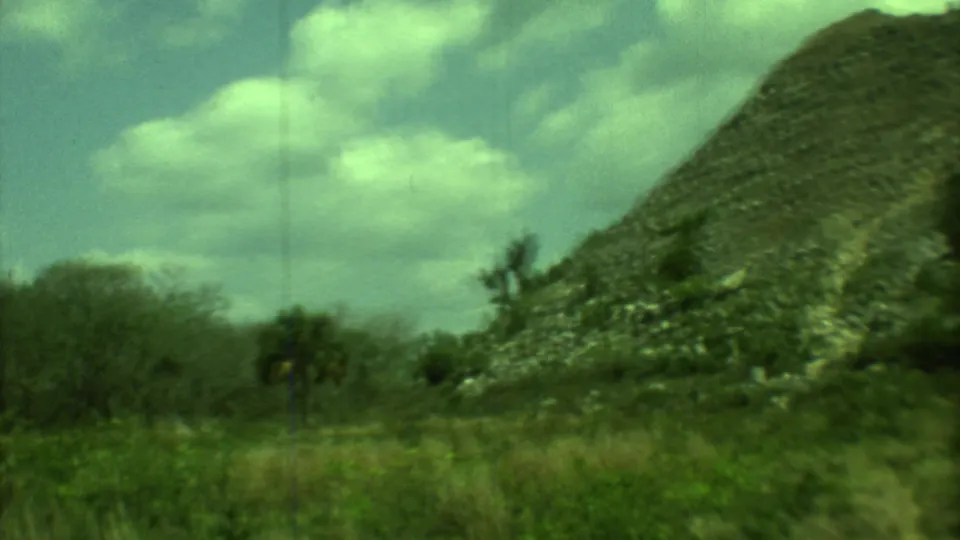 1973 Yucatan 8mm frame showing the edge of a steep mound, rural grasses, shrubs, and a bright cloud-filled sky.