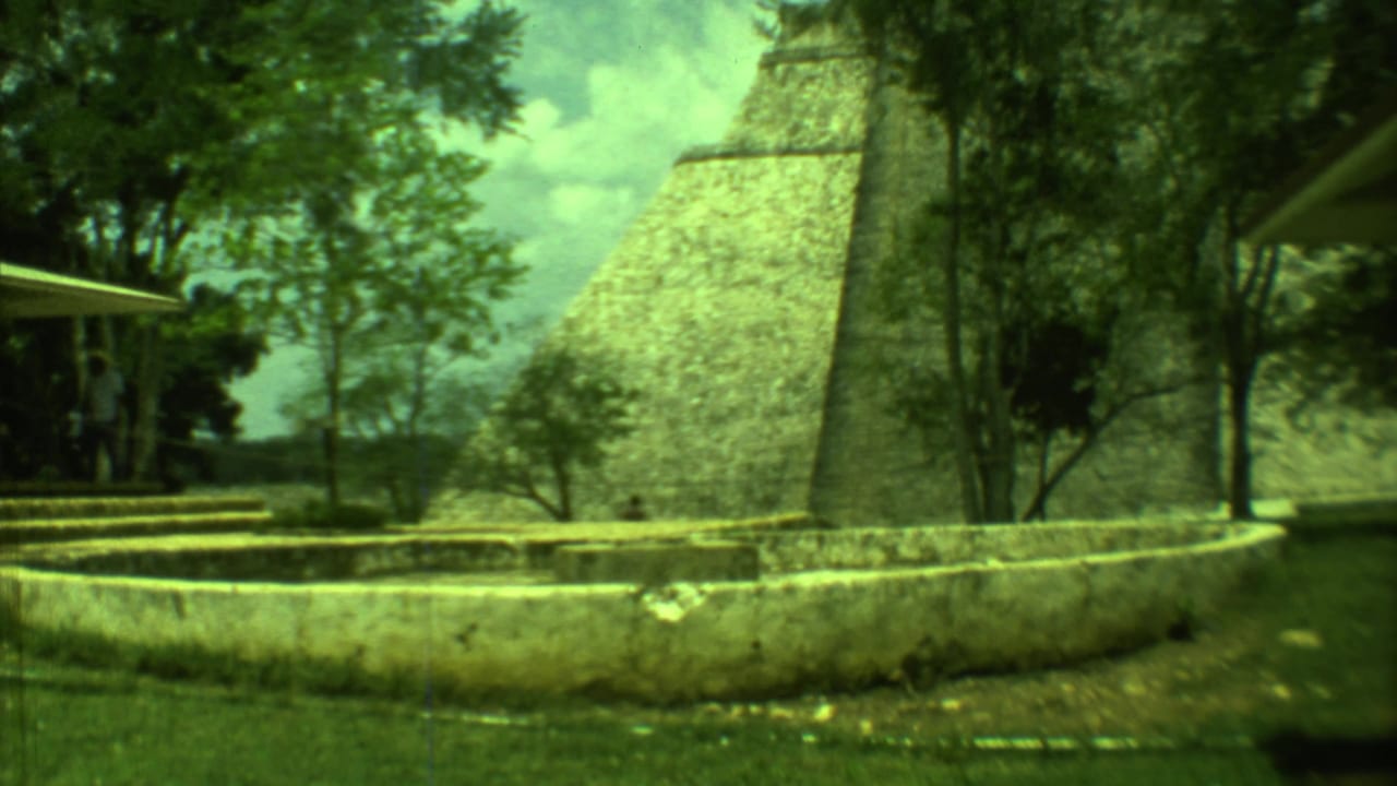 Yucatan archaeological site view with a large stone pyramid behind a circular fountain wall and surrounding trees.