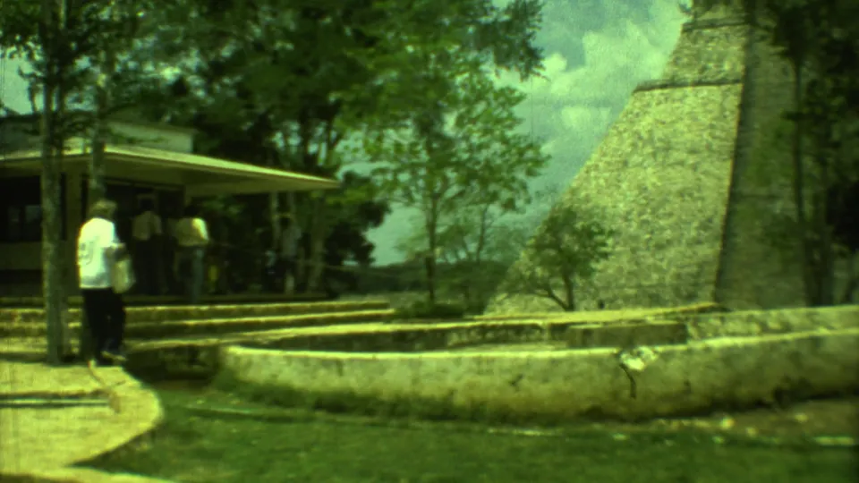 A visitor in a white shirt walks away from the camera near the fountain wall, pavilion, trees, and Yucatan pyramid.