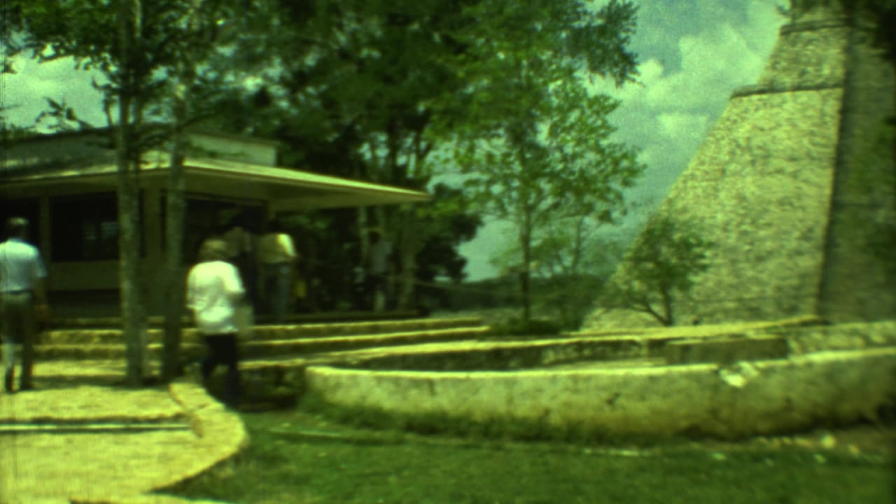 Tourists walk near a small visitor shelter and a tall stone pyramid at a ruin site.