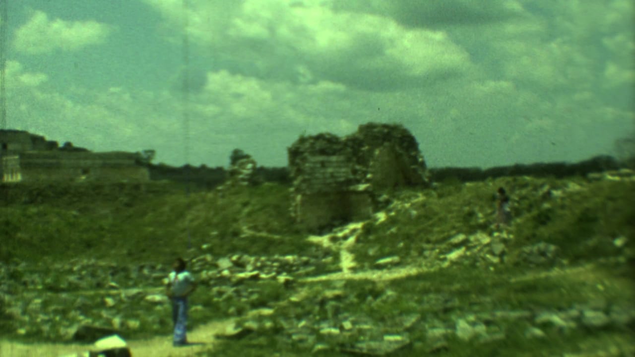 Wide view of grassy ruins with stone walls, a footpath, and a person standing in the foreground.