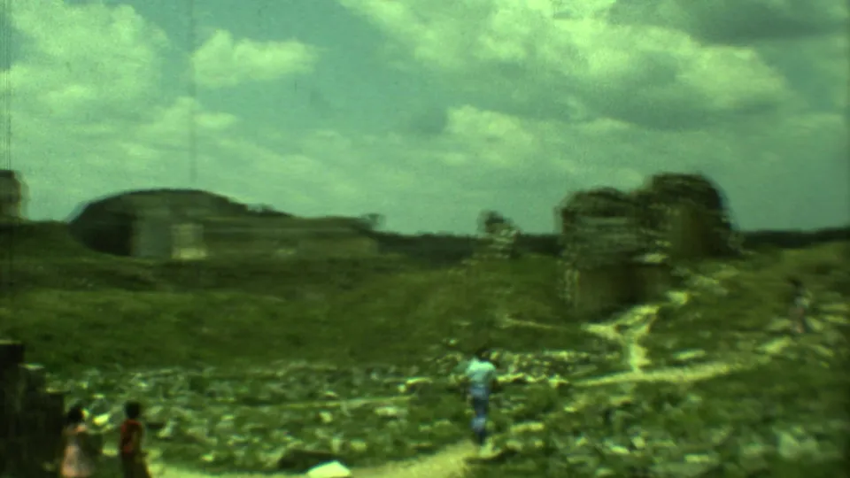 1973 Yucatan home-movie frame showing visitors dispersed across grassy Mayan ruins with stone remains and footpaths.