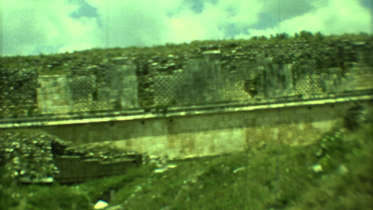 Weathered stone ruin wall with lattice openings, shrubs, and cloudy sky.