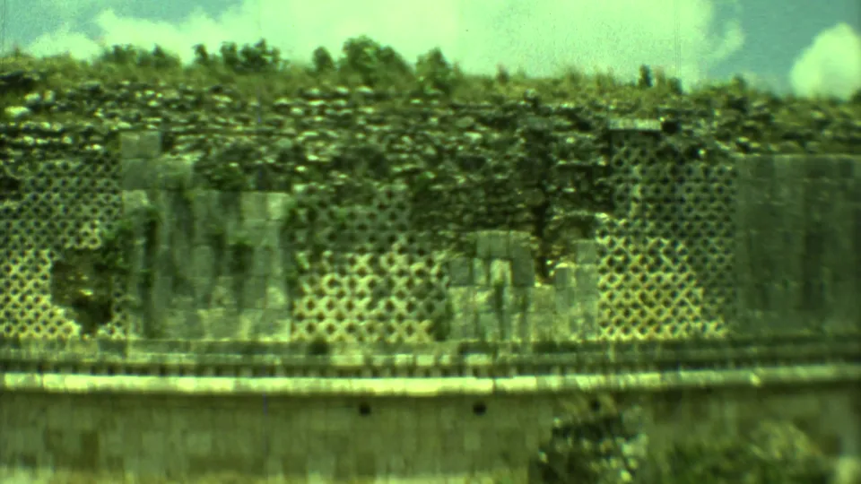Yucatan ruin wall in 1973 8mm footage, showing geometric openings, stone blocks, shrubs, and cloudy sky.