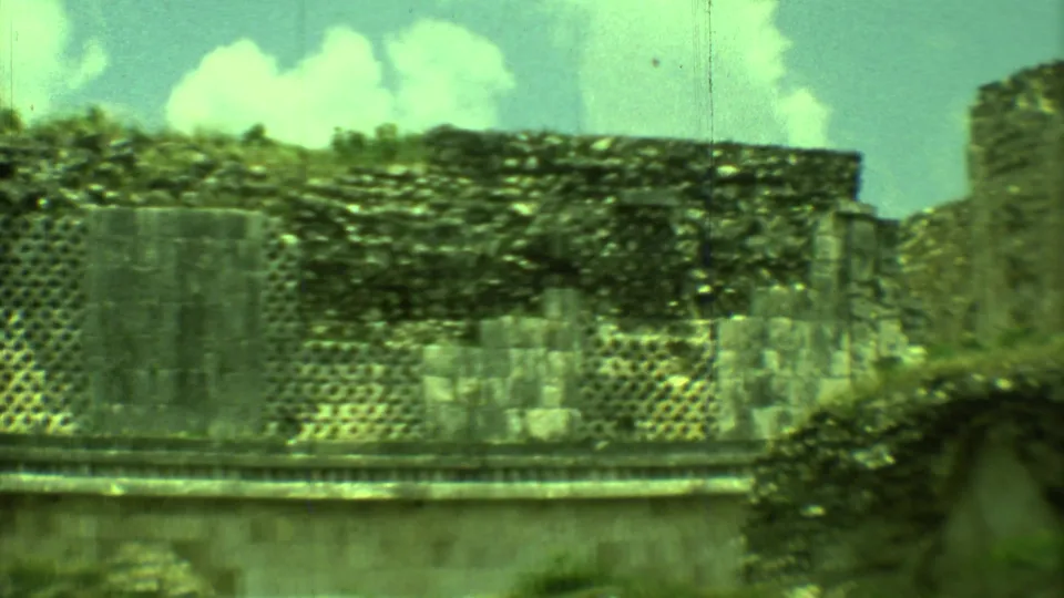 Vintage 8mm frame from Yucatan showing Mayan ruin walls, lattice-like stonework, overgrowth, and a bright clouded sky.