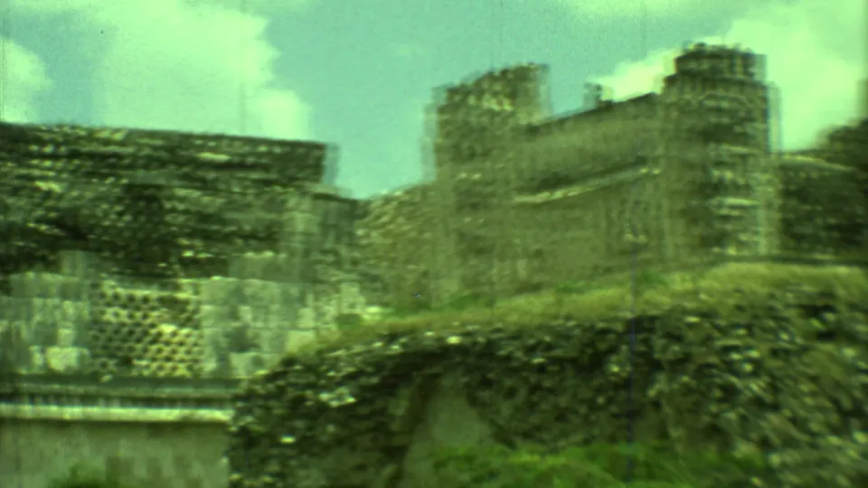 1973 Yucatan 8mm frame showing Mayan ruins with an arch-like opening, grass, dense stonework, and a taller structure to the right.