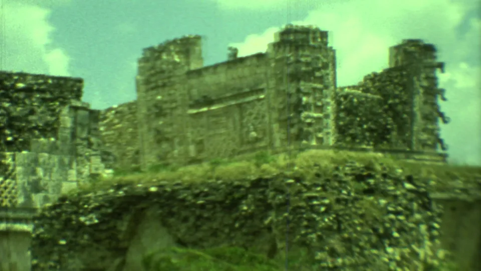 1973 Yucatan 8mm frame showing weathered Maya ruin stonework, overgrown vegetation, and a bright cloudy sky.