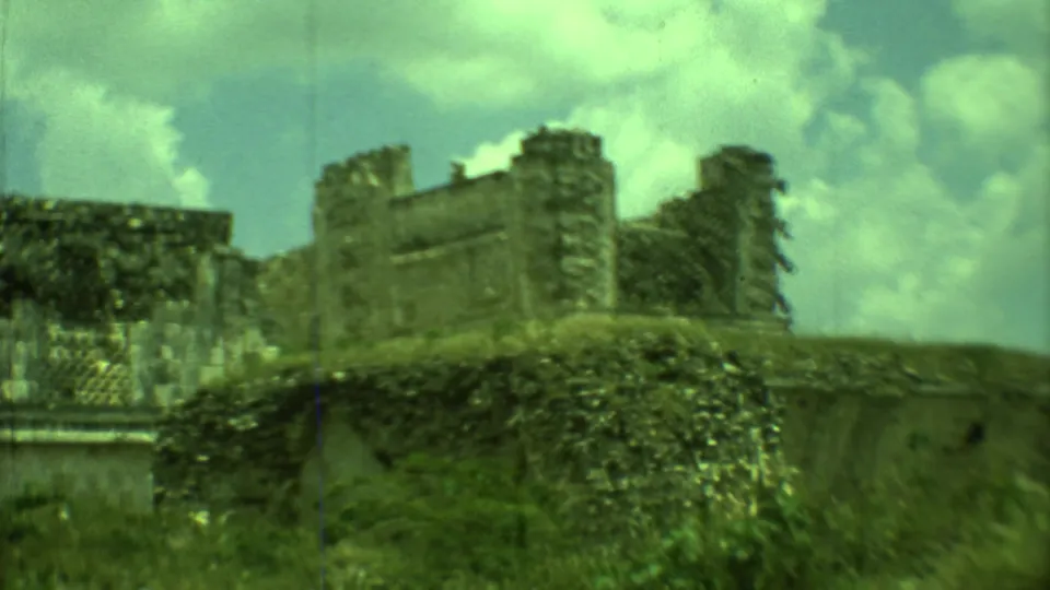 1973 Yucatan 8mm frame of an exterior Maya ruin wall, filmed from below with vegetation and bright clouds in view.