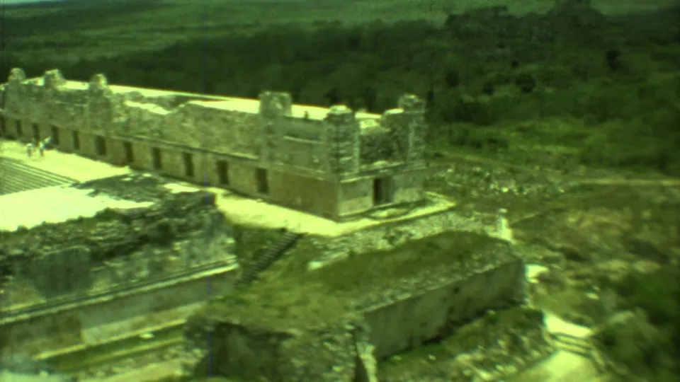 1973 Yucatan home-movie frame showing a long stone ruin complex with distant visitors on a sunlit platform.
