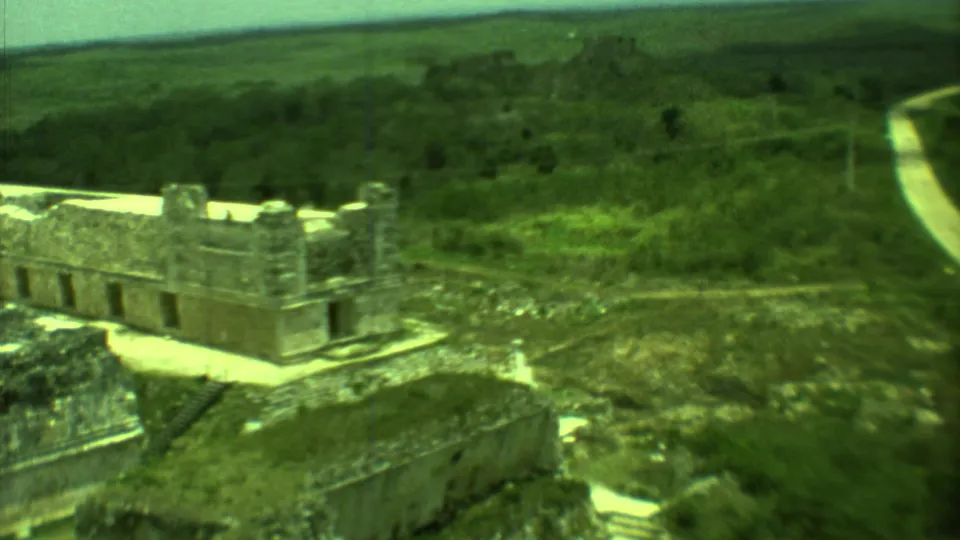 1973 Yucatan 8mm frame showing weathered ruins, surrounding vegetation, and a pale road beyond the site.