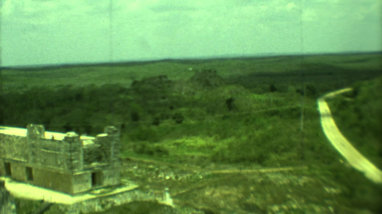 Aerial-like 1973 Yucatan view of a stone ruin near a pale road, with dense greenery and distant low hills.