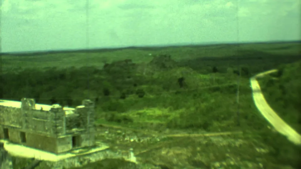 Aerial-like 1973 Yucatan view of a stone ruin near a pale road, with dense greenery and distant low hills.