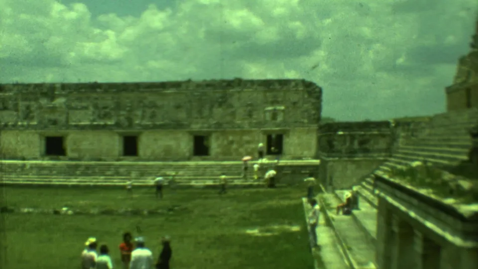 1973 home-movie frame showing a stone ruin facade, steps, and small tourist groups at a Yucatan archaeological site.