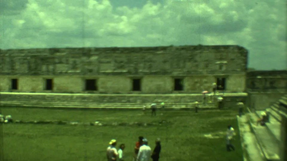 Vintage 8mm frame of tourists viewing a long Mayan ruin structure across a grassy courtyard in Yucatan, Mexico.