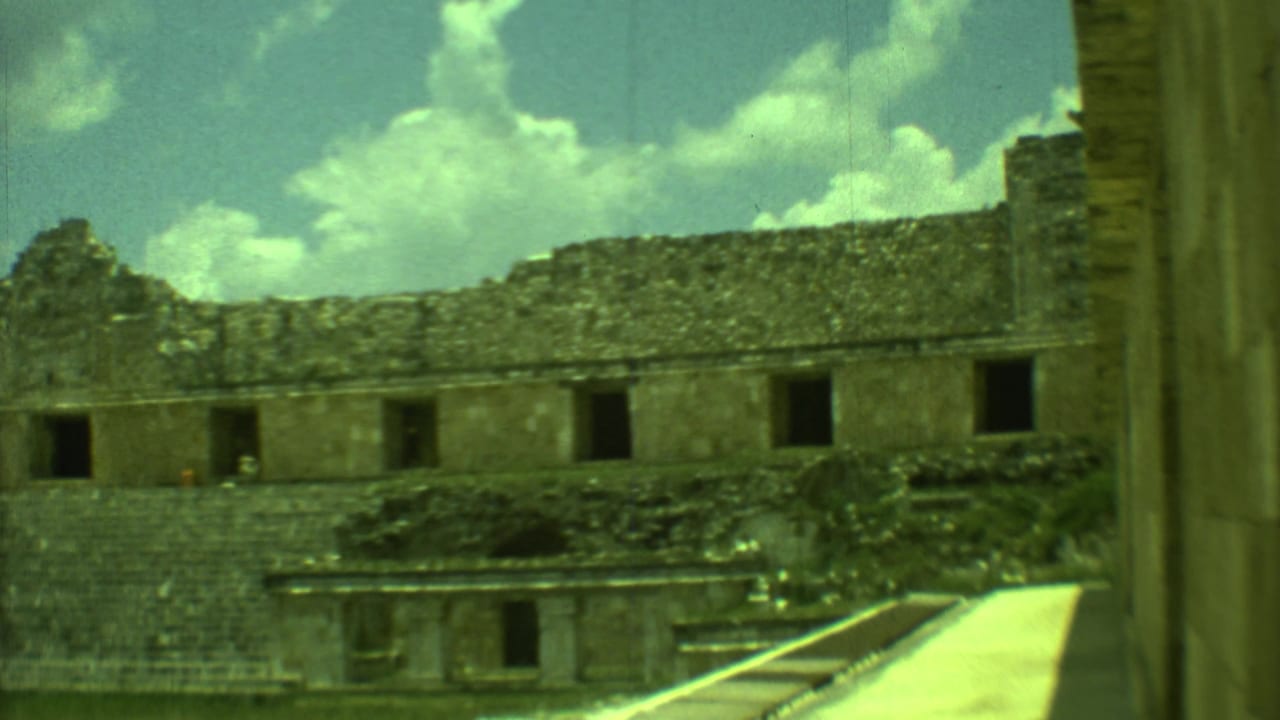 Stone ruin wall with dark doorways, stepped base, walkway, grass, and clouds in the sky.