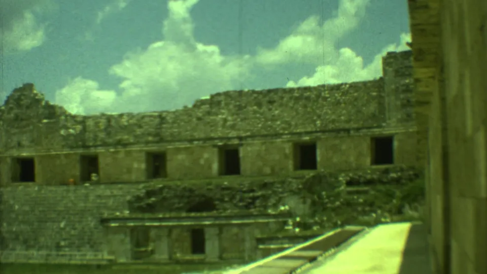 Yucatan ruins in 1973, showing a low stone structure, dark openings, tiered masonry, grass, a pale walkway, and white clouds.