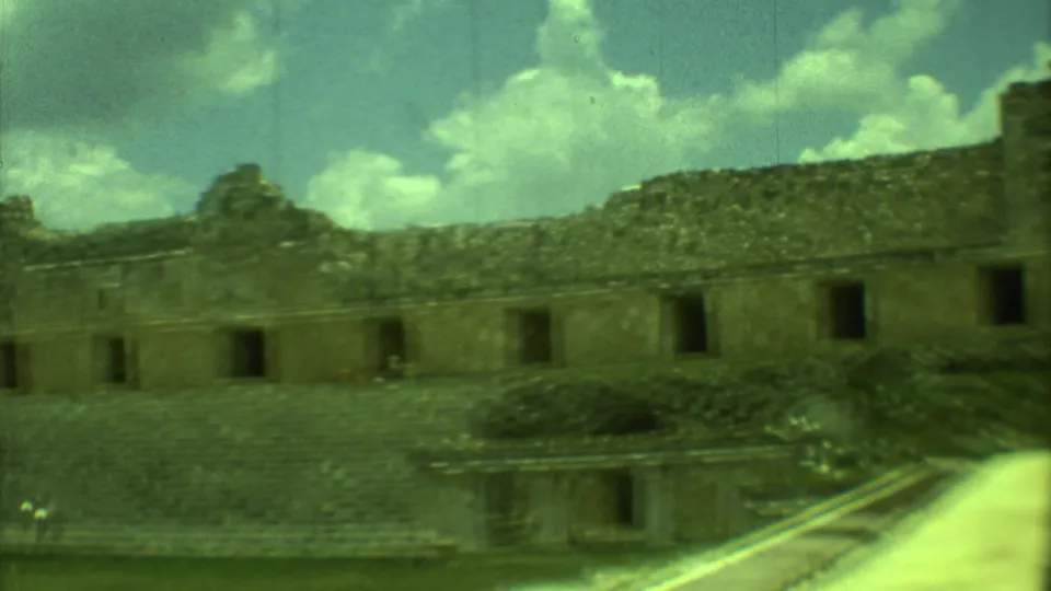 Soft 8mm frame of Yucatan ruins, with rough stone walls, dark rectangular openings, a stepped platform, grass, and bright sky.