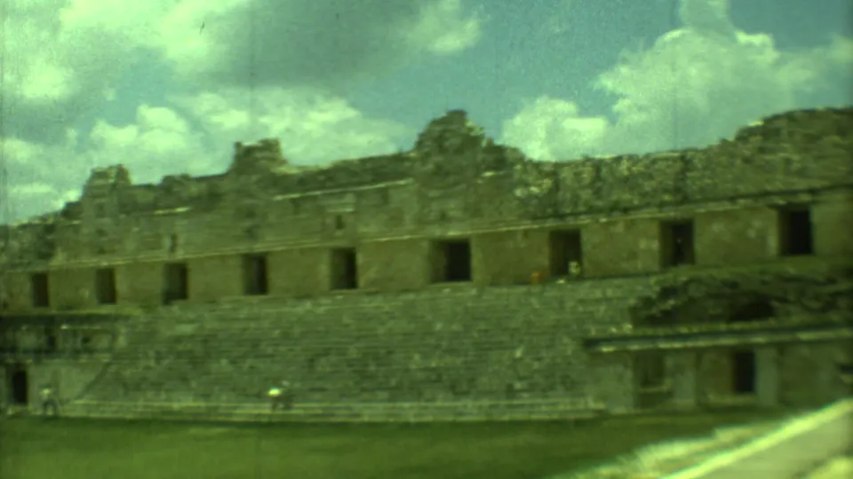 Wide 1973 Yucatan ruins view with tiered stone steps, roofless masonry, dark openings, grass, a walkway, and distant visitors.