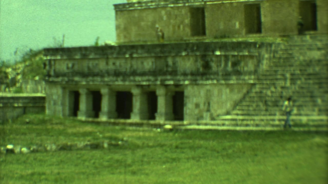 Stone building with columns and steps, seen across grass with a distant person at right.