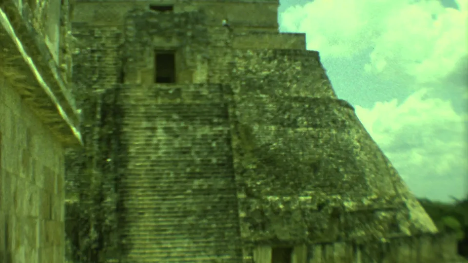 1973 Yucatan 8mm view of a stepped stone pyramid and adjacent masonry wall under a bright clouded sky.