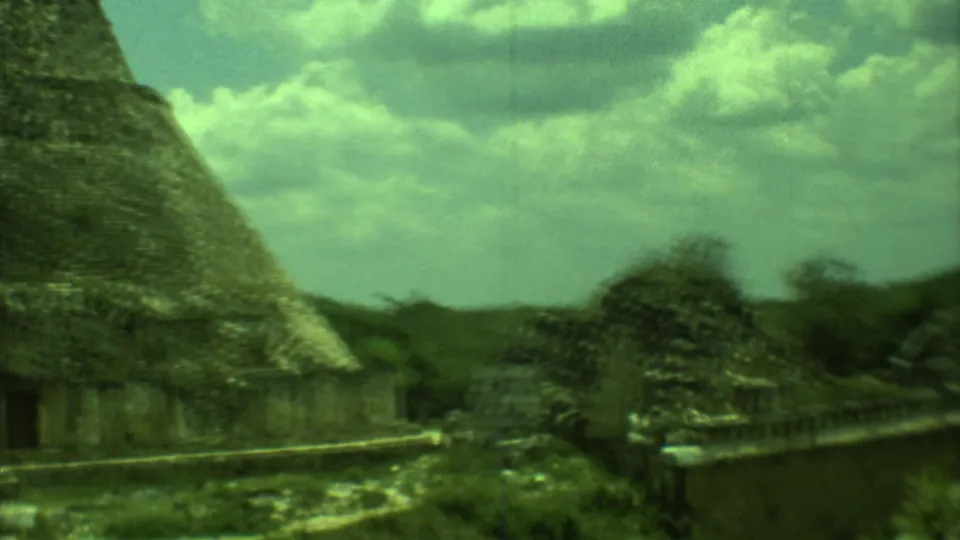 1973 8mm view of Mayan ruins in Yucatan, showing a stone structure, pointed roof form, vegetation, and cloud cover.