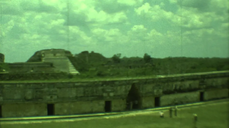 8mm home-movie frame of Yucatan ruins, with weathered masonry, a stepped structure, open lawn, and distant vegetation.