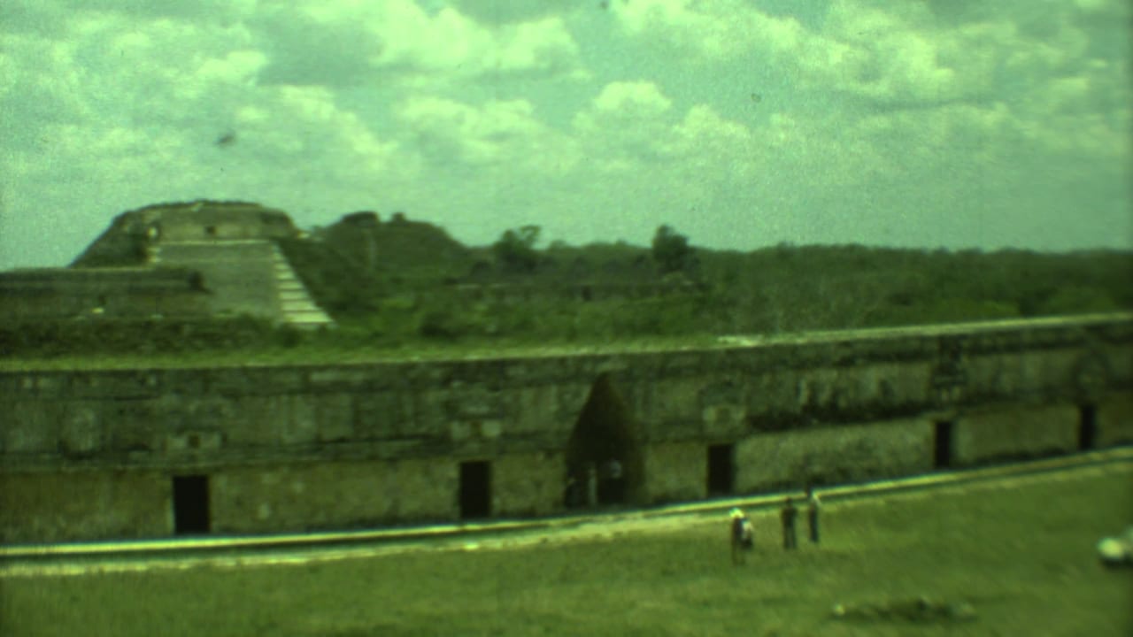 1973 8mm view in Yucatan, Mexico, showing a long stone structure, a distant pyramid form, grass, and visitors walking nearby.