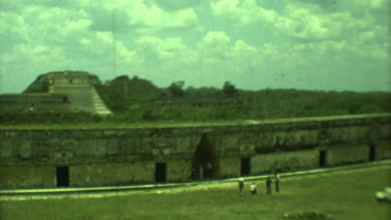 Stone ruin with dark openings, distant stepped structure, grassy area, cloudy sky, and small people.