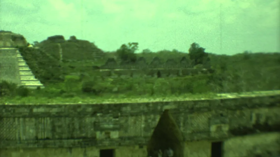 Soft 1973 8mm view of Yucatan ruins, showing weathered stone walls, a stepped pyramid edge, grass, trees, and a dark opening.