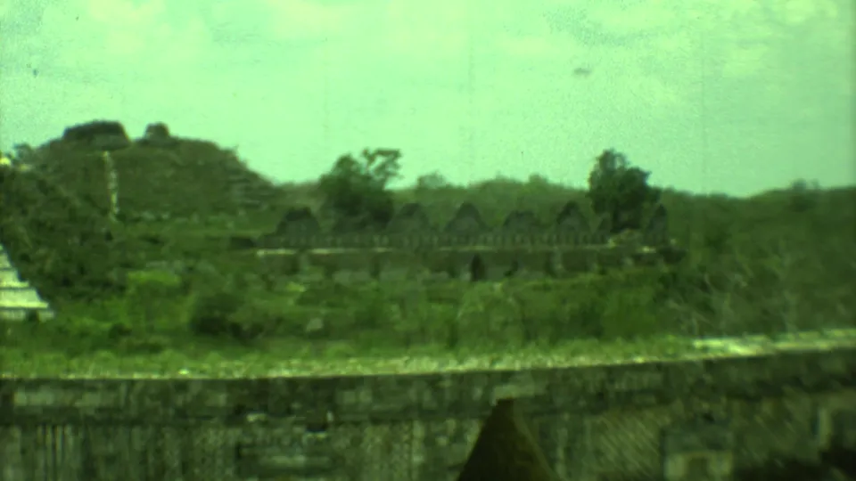 Distant 8mm frame from Yucatan, Mexico, with a pyramid-like ruin, low stone wall, grassy site, trees, and a dark doorway.