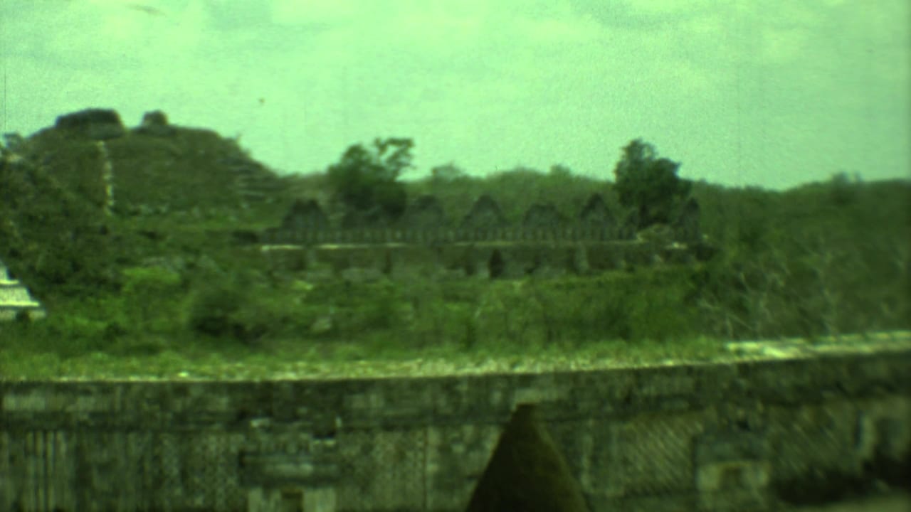1973 8mm view of Yucatan stone ruins seen from a distance, with vegetation and a cloudy sky.