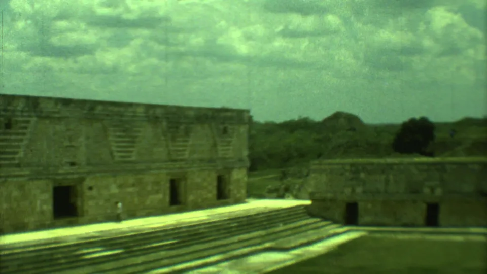 1973 Yucatan frame showing the ruin complex, stepped masonry, open courtyard, surrounding vegetation, and clouded sky.