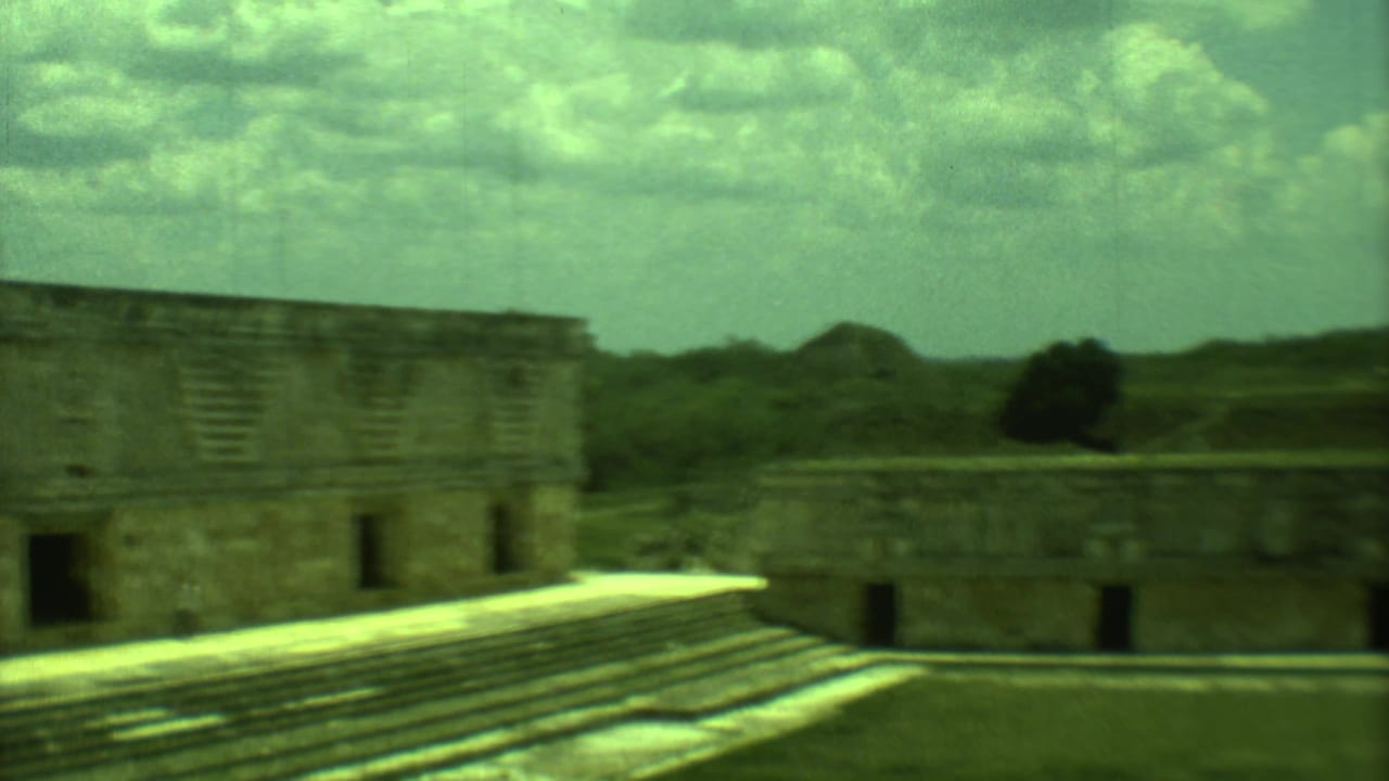 1973 8mm view of Mayan stone ruins in Yucatan, with stepped platforms, rectangular structures, and a clouded sky.
