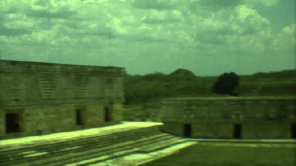 1973 8mm view of Mayan stone ruins in Yucatan, with stepped platforms, rectangular structures, and a clouded sky.